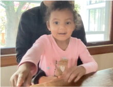 Cute child with curly brown hair sitting on a parent's lap, participating in one of the lab's Zoom looking-time studies. Shared with permission.